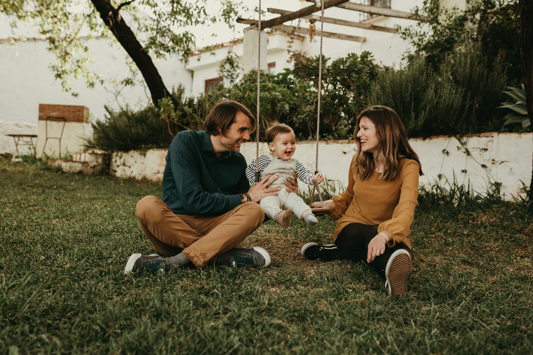 Vater und Mutter schaukeln ihr Baby, das auf dem grünen Gras im Park sitzt. Das Kind lacht glücklich.